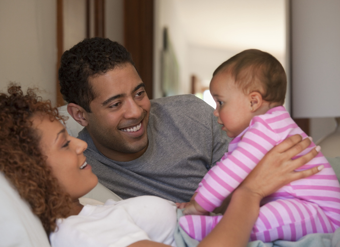 A smiling couple sitting at home with their baby daughter.
